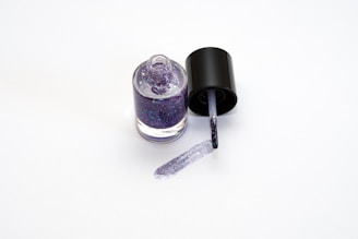 A bottle of glittery purple nail polish with the cap off and a brush resting on the surface, leaving a trail of nail polish.