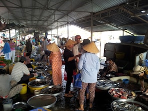 A bustling indoor market with various people, some wearing conical hats, engaging in the buying and selling of fish and other goods. Numerous bowls and containers filled with fish and seafood are spread out on the ground. The market is sheltered by a metal roof and features a variety of vendors and customers moving about.