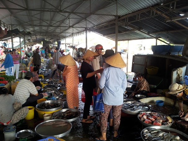 A bustling indoor market with various people, some wearing conical hats, engaging in the buying and selling of fish and other goods. Numerous bowls and containers filled with fish and seafood are spread out on the ground. The market is sheltered by a metal roof and features a variety of vendors and customers moving about.