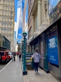 A busy urban street scene with pedestrians walking along the sidewalk. The image features tall buildings on either side, with one prominent glass skyscraper in the background. A subway entrance is visible with people heading down the stairs. A bright red vehicle is parked on the street and retail spaces with lease signs are visible.