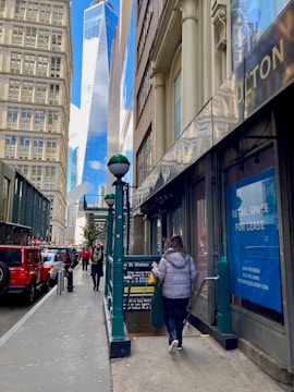 A busy urban street scene with pedestrians walking along the sidewalk. The image features tall buildings on either side, with one prominent glass skyscraper in the background. A subway entrance is visible with people heading down the stairs. A bright red vehicle is parked on the street and retail spaces with lease signs are visible.