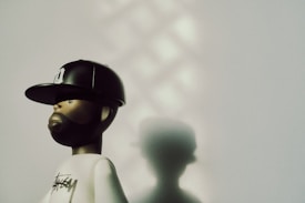 A stylized figurine with a beard and a baseball cap stands against a light wall. The figure wears a white shirt with a logo on it. Soft shadows and patterns are cast on the wall, creating an artistic and moody atmosphere.