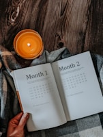 An open planner displaying Month 1 and Month 2 pages is placed on a plaid fabric, accompanied by a lit orange candle on a dark wooden surface. A hand is holding the edge of the planner.