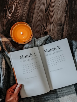 An open planner displaying Month 1 and Month 2 pages is placed on a plaid fabric, accompanied by a lit orange candle on a dark wooden surface. A hand is holding the edge of the planner.