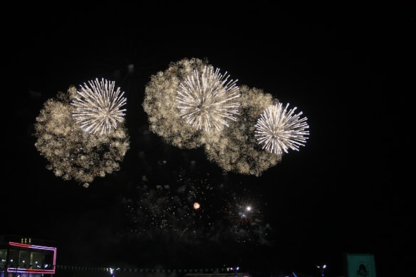 Bright and colorful fireworks exploding in a night sky. The pyrotechnic display features several large bursts that illuminate the darkness. Below, faint outlines of structures are visible, suggesting an outdoor event or celebration.