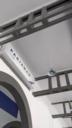 A ceiling view featuring metal beams painted in a muted gray color. A sign with the word 'Pariaman' and '+2M' is visible, suggesting a location or elevation marking. An industrial-style pendant light hangs from the beam, contributing to an understated and minimalist architectural aesthetic. The ceiling and walls are painted white, with some design elements in blue visible on the left.