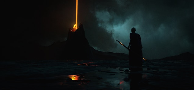 A dramatic scene unfolds with a dark, ominous sky overhead. A solitary figure stands in the foreground, silhouetted against the murky landscape, holding a staff or spear. In the distance, a mountain emits a bright, vertical beam of orange light piercing the sky. The ground appears reflective, suggesting a wet surface, amplifying the mysterious and forbidding atmosphere.