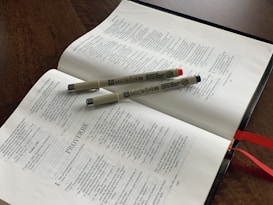 An open book is resting on a wooden table, featuring pages from the Bible, specifically the Book of Proverbs. Two Micron pens, one with a blue cap and another with a red cap, lay across the pages. Red ribbon bookmarks are visible at the edge of the book.