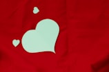 A large, light green heart cutout is placed on a vibrant red background. Two smaller heart cutouts are positioned nearby, creating a playful and romantic arrangement.