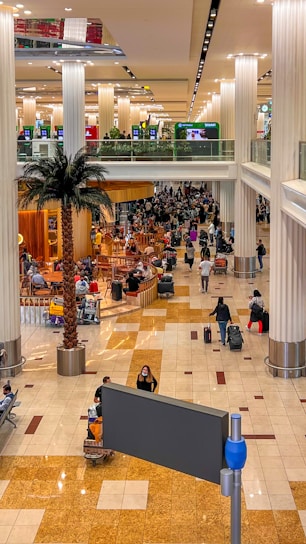 A bustling airport terminal with a large crowd of people, some standing or walking with luggage. The area is adorned with tall palm trees, many pillars, and seating arrangements around a cafe space. Bright, overhead lighting highlights the polished tiled floor, and multiple digital displays hang above the crowd.