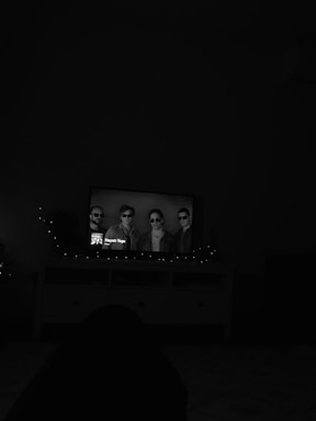 A dimly lit room features a television displaying a black-and-white image of four men wearing sunglasses. The TV is placed on a console with a dark silhouette in the foreground, possibly a person's figure. Soft ambient lighting from string lights is visible around the TV.