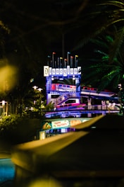 An illuminated Disneyland sign at night, surrounded by lush green foliage. Below, festive decorations with a 'Happy Holidays' banner are visible. The setting provides a festive and vibrant ambiance.