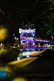 An illuminated Disneyland sign at night, surrounded by lush green foliage. Below, festive decorations with a 'Happy Holidays' banner are visible. The setting provides a festive and vibrant ambiance.