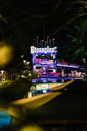 An illuminated Disneyland sign at night, surrounded by lush green foliage. Below, festive decorations with a 'Happy Holidays' banner are visible. The setting provides a festive and vibrant ambiance.