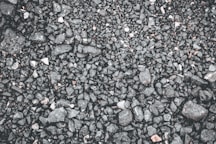 Close-up view of a gravel surface, consisting of small and medium-sized stones ranging in color from dark gray to black. The texture is uneven and rugged, with a few scattered lighter stones.