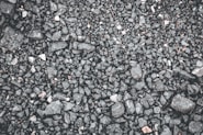 Close-up view of a gravel surface, consisting of small and medium-sized stones ranging in color from dark gray to black. The texture is uneven and rugged, with a few scattered lighter stones.