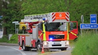 A red and yellow fire truck is positioned on a road near some grass and shrubbery, with signs pointing towards Berlin and Braunschweig. The truck is equipped with a ladder and marked with 'Feuerwehr' indicating it's part of the fire brigade. Traffic cones are placed near the truck.
