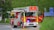 A red and yellow fire truck is positioned on a road near some grass and shrubbery, with signs pointing towards Berlin and Braunschweig. The truck is equipped with a ladder and marked with 'Feuerwehr' indicating it's part of the fire brigade. Traffic cones are placed near the truck.