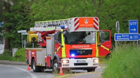 A red and yellow fire truck is positioned on a road near some grass and shrubbery, with signs pointing towards Berlin and Braunschweig. The truck is equipped with a ladder and marked with 'Feuerwehr' indicating it's part of the fire brigade. Traffic cones are placed near the truck.