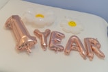 Metallic gold balloons spell out '1 YEAR' and are accompanied by two inflatable white flowers with yellow centers on a white surface, possibly a bed or table.