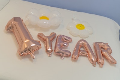 Metallic gold balloons spell out '1 YEAR' and are accompanied by two inflatable white flowers with yellow centers on a white surface, possibly a bed or table.