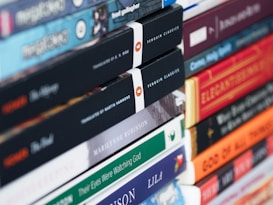 A collection of various books stacked horizontally and vertically. The spines show a range of titles and authors, with some books belonging to the Penguin Classics series. The books are stacked neatly, suggesting a well-organized library or personal collection.