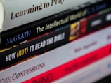 A close-up view of a staggered stack of books with varying titles and colors, some with bold or italicized text. The books appear to focus on religious or philosophical topics.