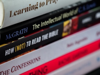 A close-up view of a staggered stack of books with varying titles and colors, some with bold or italicized text. The books appear to focus on religious or philosophical topics.