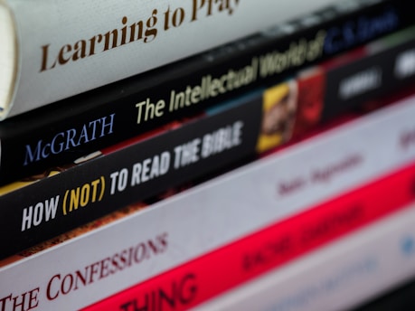 A close-up view of a staggered stack of books with varying titles and colors, some with bold or italicized text. The books appear to focus on religious or philosophical topics.