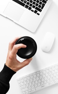 A person's hand is holding a black camera lens cap above a white table surface. Nearby, there is an open laptop, a wireless computer mouse, and a wireless keyboard, all in white. The setting appears clean and minimalistic.