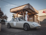 A classic car is parked in front of a vintage gas station with retro signage, including brands like Shell and BP. The station has a rustic, historic appearance with a corrugated metal roof and worn-out facade. The car is sleek and well-maintained, highlighting its vintage design.