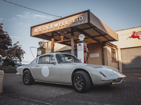 A classic car is parked in front of a vintage gas station with retro signage, including brands like Shell and BP. The station has a rustic, historic appearance with a corrugated metal roof and worn-out facade. The car is sleek and well-maintained, highlighting its vintage design.