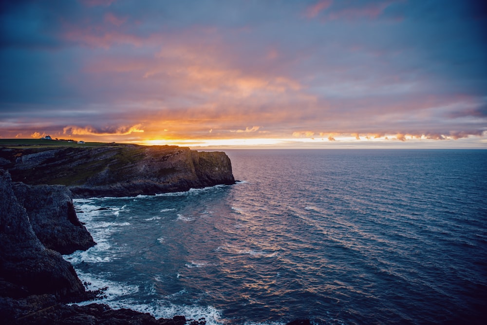 the sun is setting over the ocean near a cliff