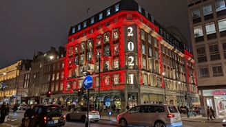 A building with festive decorations is illuminated with red lights and the year 2021 displayed prominently. The streets below are busy with cars and pedestrians. The building has windows decorated with Christmas themes, suggesting a holiday season atmosphere.