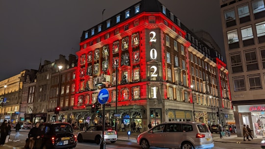 A building with festive decorations is illuminated with red lights and the year 2021 displayed prominently. The streets below are busy with cars and pedestrians. The building has windows decorated with Christmas themes, suggesting a holiday season atmosphere.