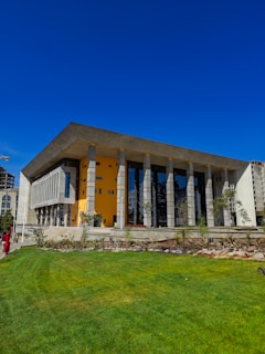 A modern building with a unique architectural design featuring tall rectangular columns, a bright yellow wall, and large glass windows. The foreground displays a well-maintained green lawn with several plants and a clear blue sky above.