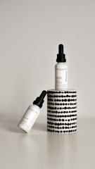 Two dropper bottles labeled ‘Revolution’ with the product name ‘Intense Acid Peel’ are displayed against a minimalist background. One bottle is upright on a cylindrical stand with a black and white dotted pattern, while the other bottle is leaning diagonally at the base.