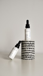 Two dropper bottles labeled ‘Revolution’ with the product name ‘Intense Acid Peel’ are displayed against a minimalist background. One bottle is upright on a cylindrical stand with a black and white dotted pattern, while the other bottle is leaning diagonally at the base.