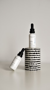Two dropper bottles labeled ‘Revolution’ with the product name ‘Intense Acid Peel’ are displayed against a minimalist background. One bottle is upright on a cylindrical stand with a black and white dotted pattern, while the other bottle is leaning diagonally at the base.