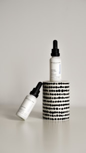 Two dropper bottles labeled ‘Revolution’ with the product name ‘Intense Acid Peel’ are displayed against a minimalist background. One bottle is upright on a cylindrical stand with a black and white dotted pattern, while the other bottle is leaning diagonally at the base.
