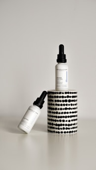 Two dropper bottles labeled ‘Revolution’ with the product name ‘Intense Acid Peel’ are displayed against a minimalist background. One bottle is upright on a cylindrical stand with a black and white dotted pattern, while the other bottle is leaning diagonally at the base.