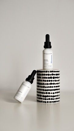 Two dropper bottles labeled ‘Revolution’ with the product name ‘Intense Acid Peel’ are displayed against a minimalist background. One bottle is upright on a cylindrical stand with a black and white dotted pattern, while the other bottle is leaning diagonally at the base.