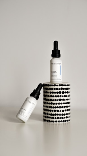 Two dropper bottles labeled ‘Revolution’ with the product name ‘Intense Acid Peel’ are displayed against a minimalist background. One bottle is upright on a cylindrical stand with a black and white dotted pattern, while the other bottle is leaning diagonally at the base.