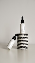 Two dropper bottles labeled ‘Revolution’ with the product name ‘Intense Acid Peel’ are displayed against a minimalist background. One bottle is upright on a cylindrical stand with a black and white dotted pattern, while the other bottle is leaning diagonally at the base.