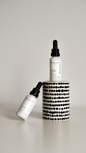 Two dropper bottles labeled ‘Revolution’ with the product name ‘Intense Acid Peel’ are displayed against a minimalist background. One bottle is upright on a cylindrical stand with a black and white dotted pattern, while the other bottle is leaning diagonally at the base.