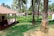 A landscaped garden with neatly trimmed grass, a variety of palm trees, and a traditional house with a tiled roof and a porch adorned with hanging plants. The setting is lush and verdant, suggesting a tropical environment.