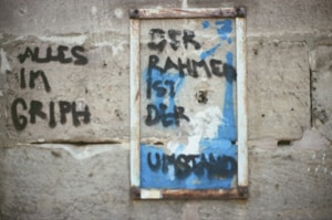 Graffiti text in black paint on a rough, beige concrete wall. The text appears in two sections, with one phrase partially framed within a worn, painted blue rectangle.