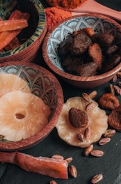 An assortment of dried fruits and nuts is arranged in decorative bowls and scattered on a dark surface. The bowls contain dried apricots, pistachios, and slices of dried pineapple. The intricate patterns on the bowls add an artistic touch to the rustic presentation.