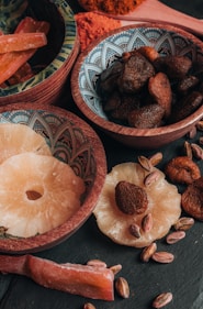 An assortment of dried fruits and nuts is arranged in decorative bowls and scattered on a dark surface. The bowls contain dried apricots, pistachios, and slices of dried pineapple. The intricate patterns on the bowls add an artistic touch to the rustic presentation.