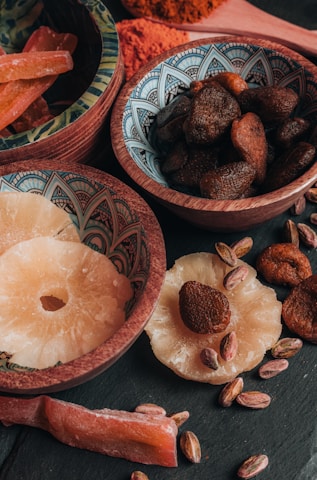 An assortment of dried fruits and nuts is arranged in decorative bowls and scattered on a dark surface. The bowls contain dried apricots, pistachios, and slices of dried pineapple. The intricate patterns on the bowls add an artistic touch to the rustic presentation.